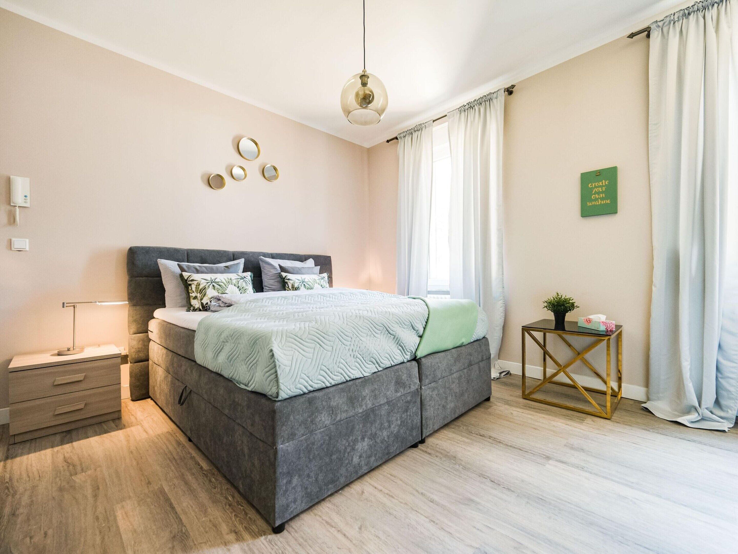 Een moderne slaapkamer met een grijs gestoffeerd bed, een nachtkastje met een lamp, een klein bijzettafeltje met een plant en een spiegel aan de muur.