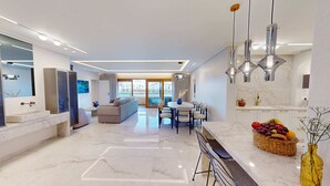 Una cocina moderna con encimera de mármol, un comedor con mesa y sillas redondas, y una sala de estar con sofá y ventana grande.
