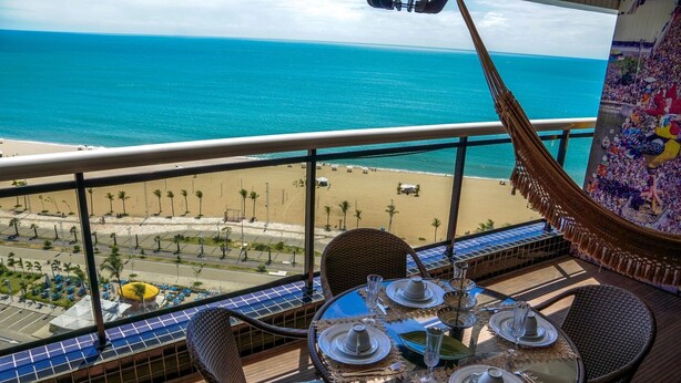 A balcony with a table set for dining, overlooking a beach and ocean.