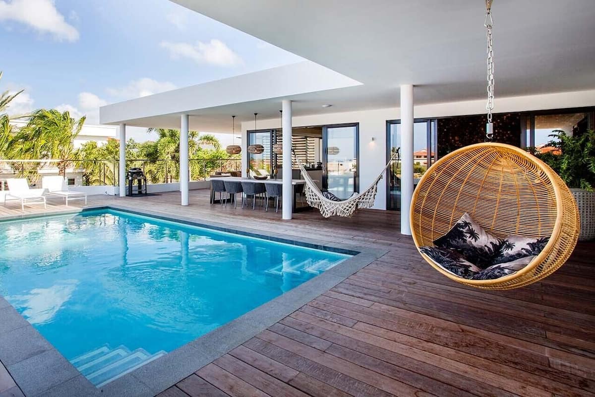 Un área de piscina exterior moderna con terraza de madera, una silla colgante y un comedor con mobiliario de exterior.