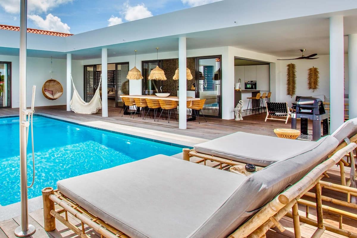 Un área de piscina moderna con sillones reclinables, un comedor y una cocina visibles a través de puertas de cristal.