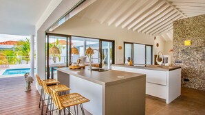 A modern kitchen with a large island, bar stools, and a view of the pool and palm trees outside. - The Villas by LXRYHOME (Jan Thiel)