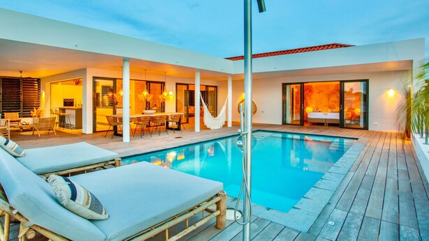 A modern outdoor pool area with lounge chairs, a hammock, and a glass-enclosed spa room. - The Villas by LXRYHOME (Jan Thiel)