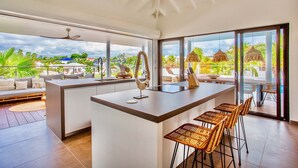 A modern kitchen with a central island, bar stools, and large windows offering a view of the outdoors. - The Villas by LXRYHOME (Jan Thiel)