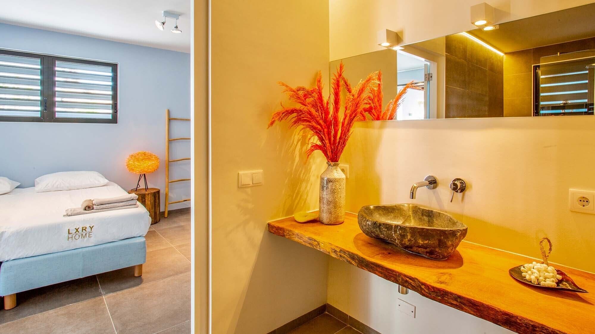A modern hotel room with a bed, a wooden vanity with a bowl sink, a vase with red feathers, and a mirror above the sink.