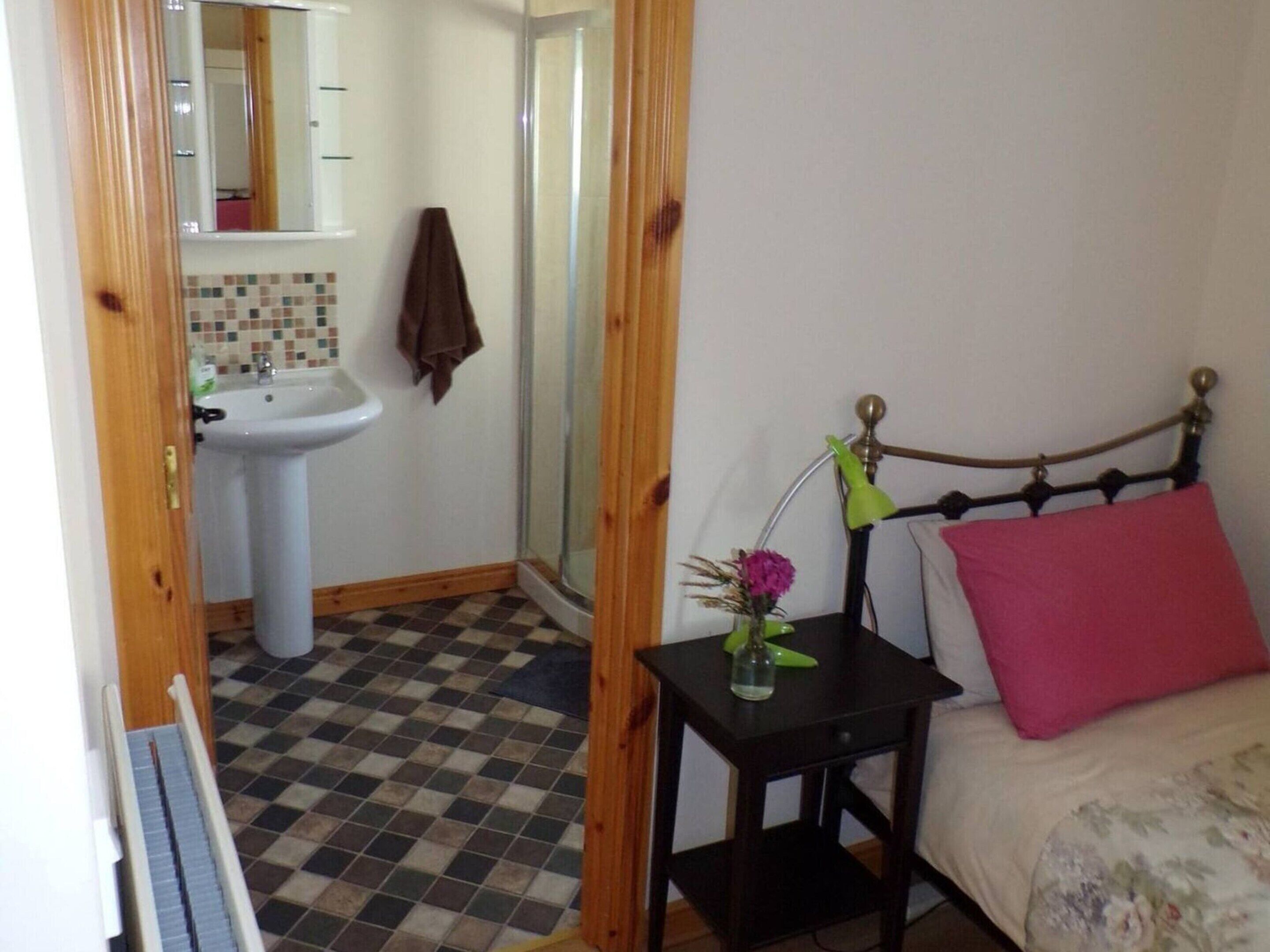 A bedroom with a bed, a bedside table with a lamp, a small table with a vase, a bathroom with a sink and mirror, and a checkered floor.