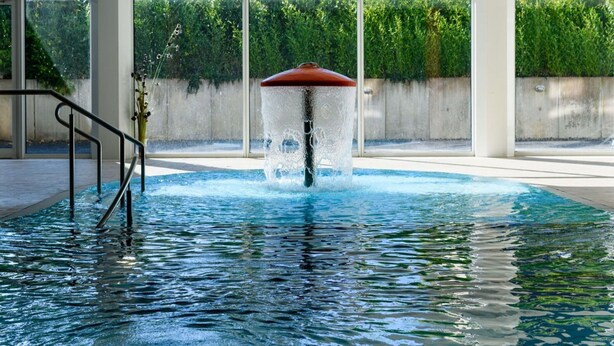 Une piscine agrémentée d’une fontaine, entourée de grandes fenêtres et d’un mur en béton.