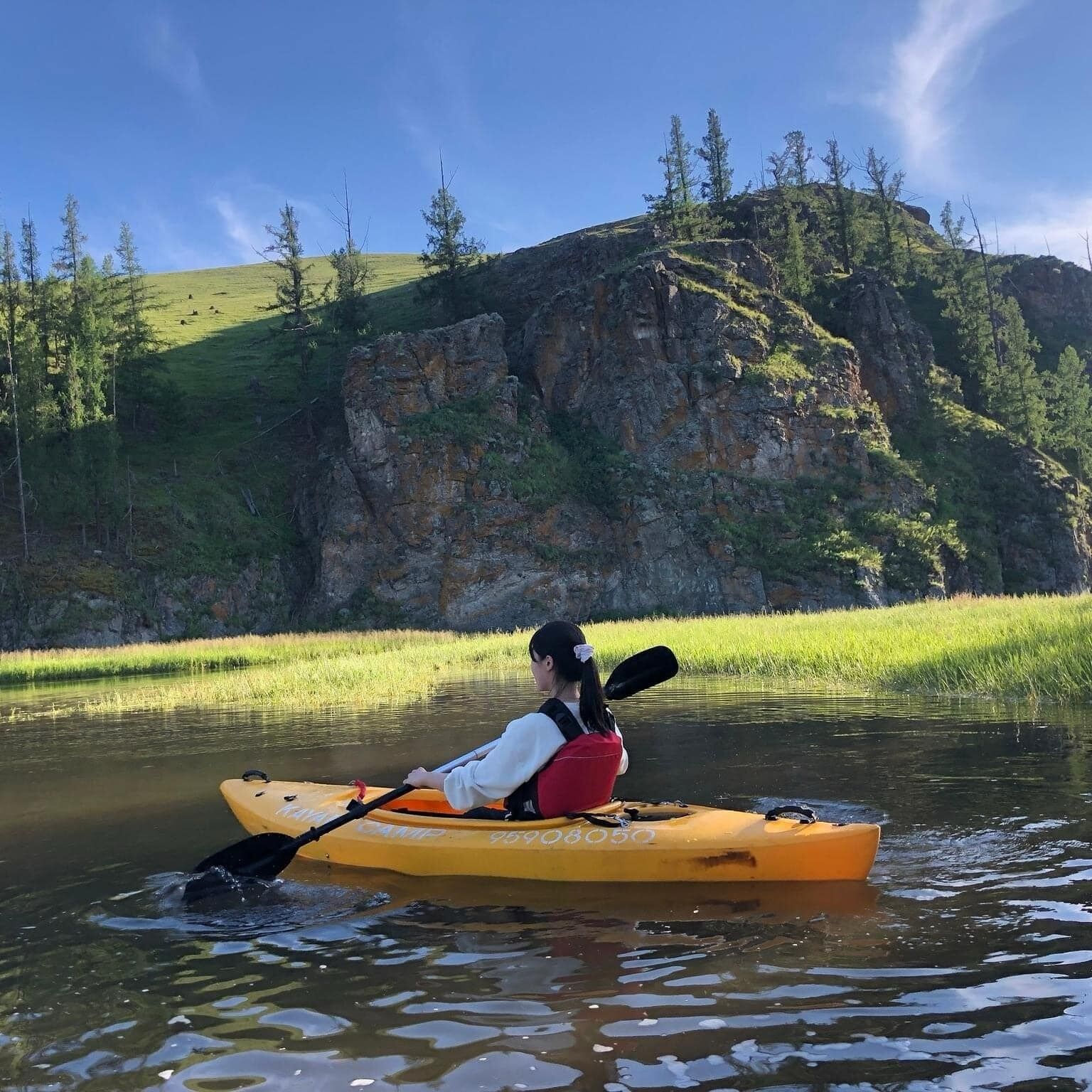 A person kayaking on a calm river surrounded by grassy banks and rocky hills.