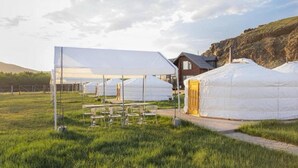 Des yourtes à la structure blanche, ressemblant à des tentes, se dressent dans un champ verdoyant, à côté d’un bâtiment en bois, sous un ciel dégagé.