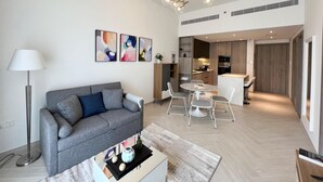 A modern living room with a grey sofa, a coffee table with decorative items, and a dining area with a round table and chairs. - Waves - Comfortable JVC Apt with Balcony (Dubai)