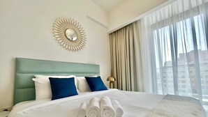 A hotel room with a large bed, a headboard, a wall-mounted mirror, a window with curtains, and a view of buildings outside. - Waves - Comfortable JVC Apt with Balcony (Dubai)