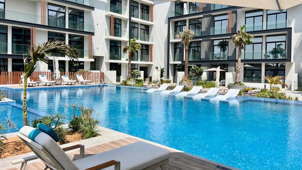 A modern hotel pool area with multiple lounge chairs, palm trees, and a clear blue pool. - Waves - Comfortable JVC Apt with Balcony (Dubai)
