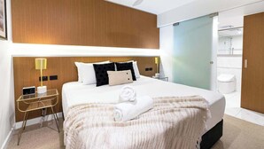 Une chambre d’hôtel avec un grand lit, des tables de chevet, un bureau, un ventilateur de plafond et une salle de bain visible à travers une porte ouverte.