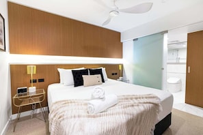 Camera d'albergo con un letto grande, comodini, una scrivania, una ventola a soffitto e un bagno visibile attraverso una porta aperta.