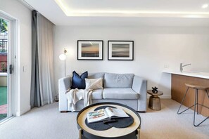 A modern living room with a grey sofa, a coffee table with a magazine, two framed pictures on the wall, and a kitchen area with a bar stool.