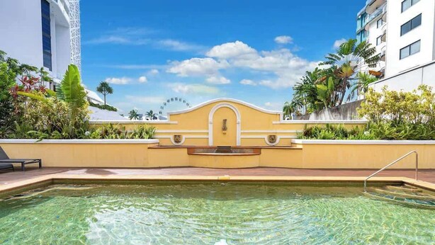 Une piscine aux eaux cristallines avec un mur jaune, un bâtiment blanc doté de balcons et des plantes tropicales.