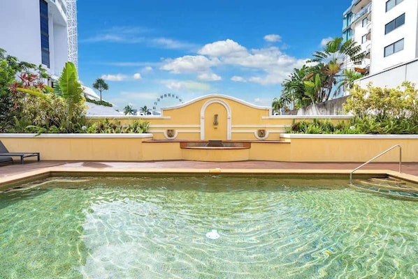 Una piscina trasparente con una parete gialla, un edificio bianco con balconi e piante tropicali.