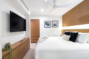 A modern hotel room with a flat-screen TV, a bed with white bedding, a wooden headboard, and a ceiling fan.