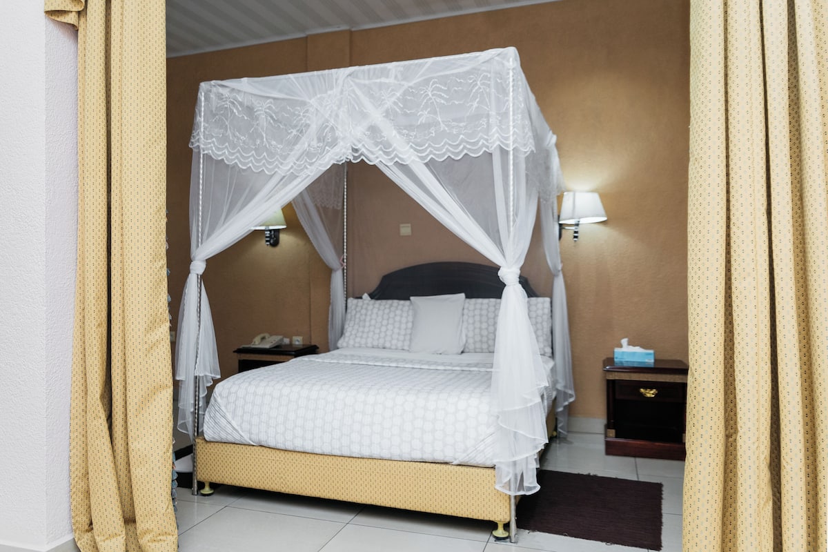 Een hemelbed met een wit beddegoed en kussens, een nachtkastje met een lamp, en een bijzettafeltje met een telefoon en zakdoekjes.