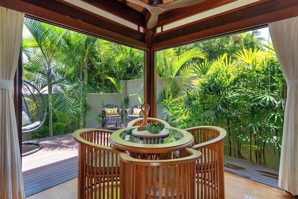 A room with a wooden table and chairs, large windows, and a view of lush greenery outside.