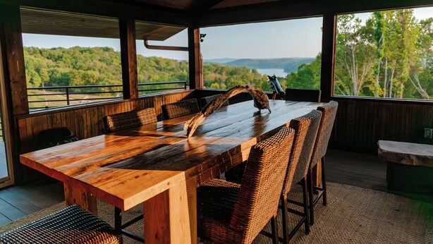 A wooden dining table with wicker chairs, large windows with a view of a forest and mountains, and a stone bench. - Spacious lakeview lodge on Lake Cumberland with WiFi, AC (Seventy Six)