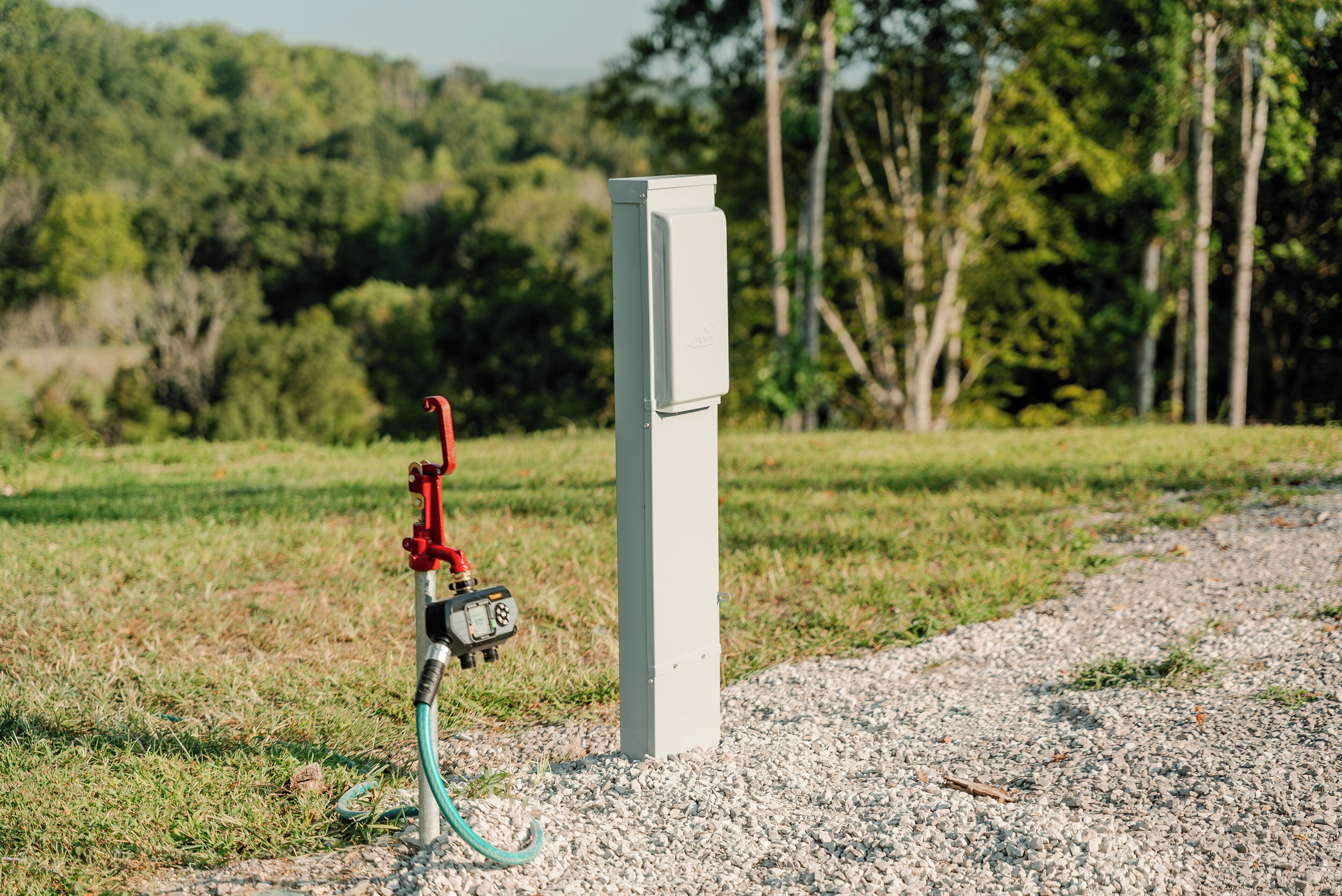 A utility box with a red fire hydrant attached to it, connected by a green hose, set in a gravel area with grass and trees in the background.