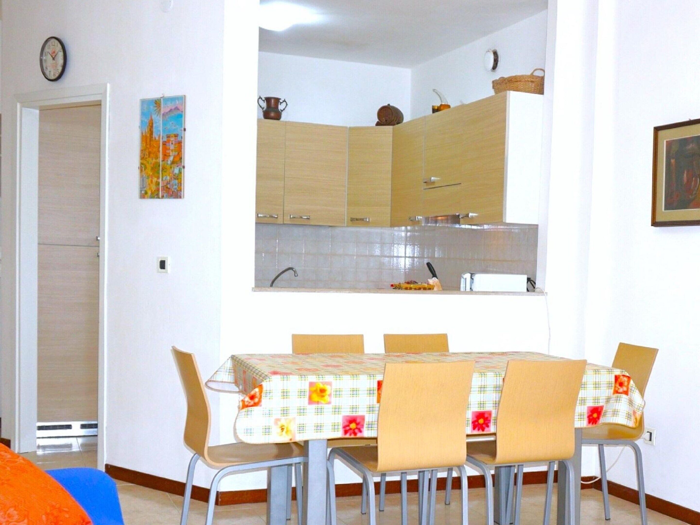 A dining area with a table set for four, surrounded by chairs. A kitchen with wooden cabinets and a tiled backsplash is visible in the background.
