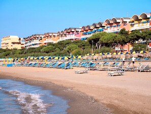 A beach with rows of blue and yellow sun loungers and umbrellas, a sandy shore, and a row of colorful buildings in the background. - Elegant Group Stay Near Caorle (Caorle)