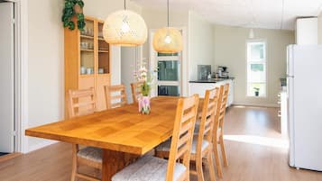 A dining area with a wooden table and chairs, a kitchen in the background, and a hanging light fixture.