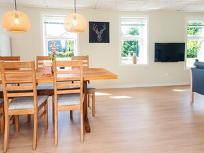 A dining area with a wooden table and chairs, two hanging lamps, a wall-mounted TV, and windows with a view of greenery. - 4 Star Holiday Home in Hovborg (Hovborg)