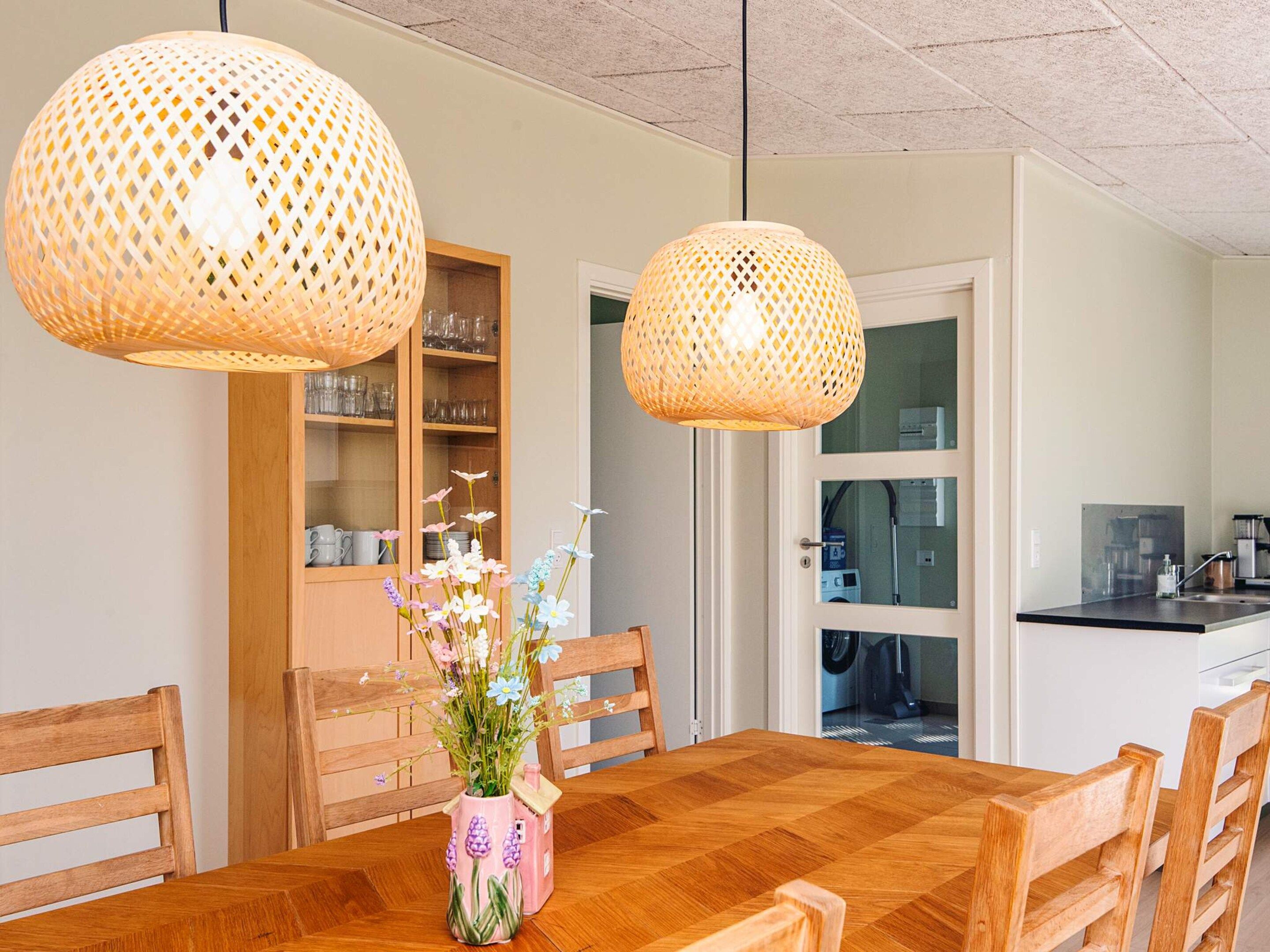 A dining area with a wooden table, chairs, a pink vase with flowers, and two wicker pendant lights.