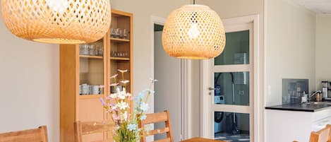 A dining area with a wooden table, chairs, a pink vase with flowers, and two wicker pendant lights.