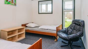 A dormitory room with a single bed, a wooden desk, a black chair, and two windows with blinds.