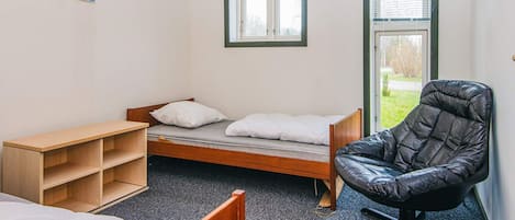 A dormitory room with a single bed, a wooden desk, a black chair, and two windows with blinds.