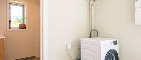 A laundry room with a front-loading washing machine, a water heater, and a window with a plant.