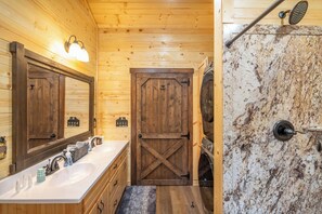 A wooden cabin bathroom with a large mirror, a sink with toiletries, a wooden door, and a shower area.