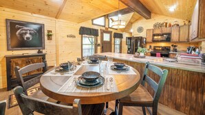 A wooden cabin interior with a dining table set for four, a kitchen with modern appliances, and a bear-themed artwork on the wall. - 🔥 Tennessee Dream | Cozy Retreat with Fire Pit + Mortal Kombat Arcade🌲 (Sevierville)
