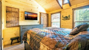 A cozy log cabin bedroom with a large bed, a dresser, and a painting of a sunset. - 🔥 Tennessee Dream | Cozy Retreat with Fire Pit + Mortal Kombat Arcade🌲 (Sevierville)