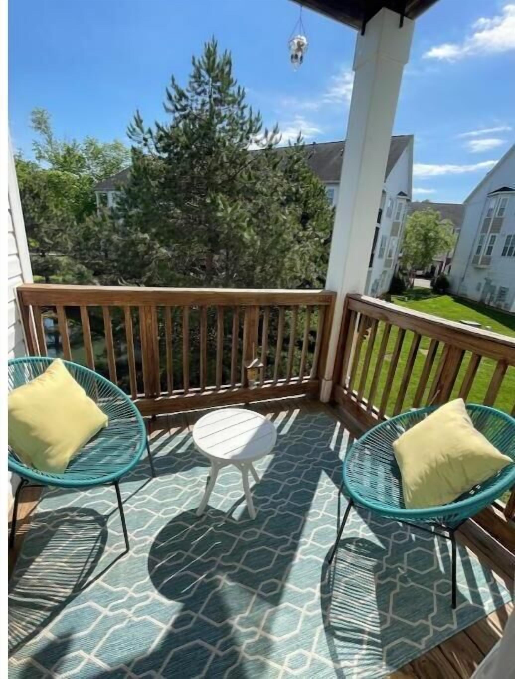 Un espacio al aire libre techado con piso de madera, dos sillas de mimbre verde con cojines amarillos y una pequeña mesa blanca.