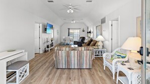 A spacious living area with a large striped sofa, a white wicker chair, and a dining table. There is a ceiling fan, a flat-screen TV mounted on the wall, and a window with curtains. - Bubba's Beach Blessing - 4BR/2BA- Pet Friendly (North Myrtle Beach)