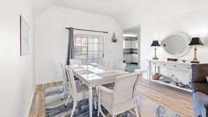 A dining area with a glass-top table and white wicker chairs, a kitchen in the background, and a blue patterned rug on the floor. - Bubba's Beach Blessing - 4BR/2BA- Pet Friendly (North Myrtle Beach)