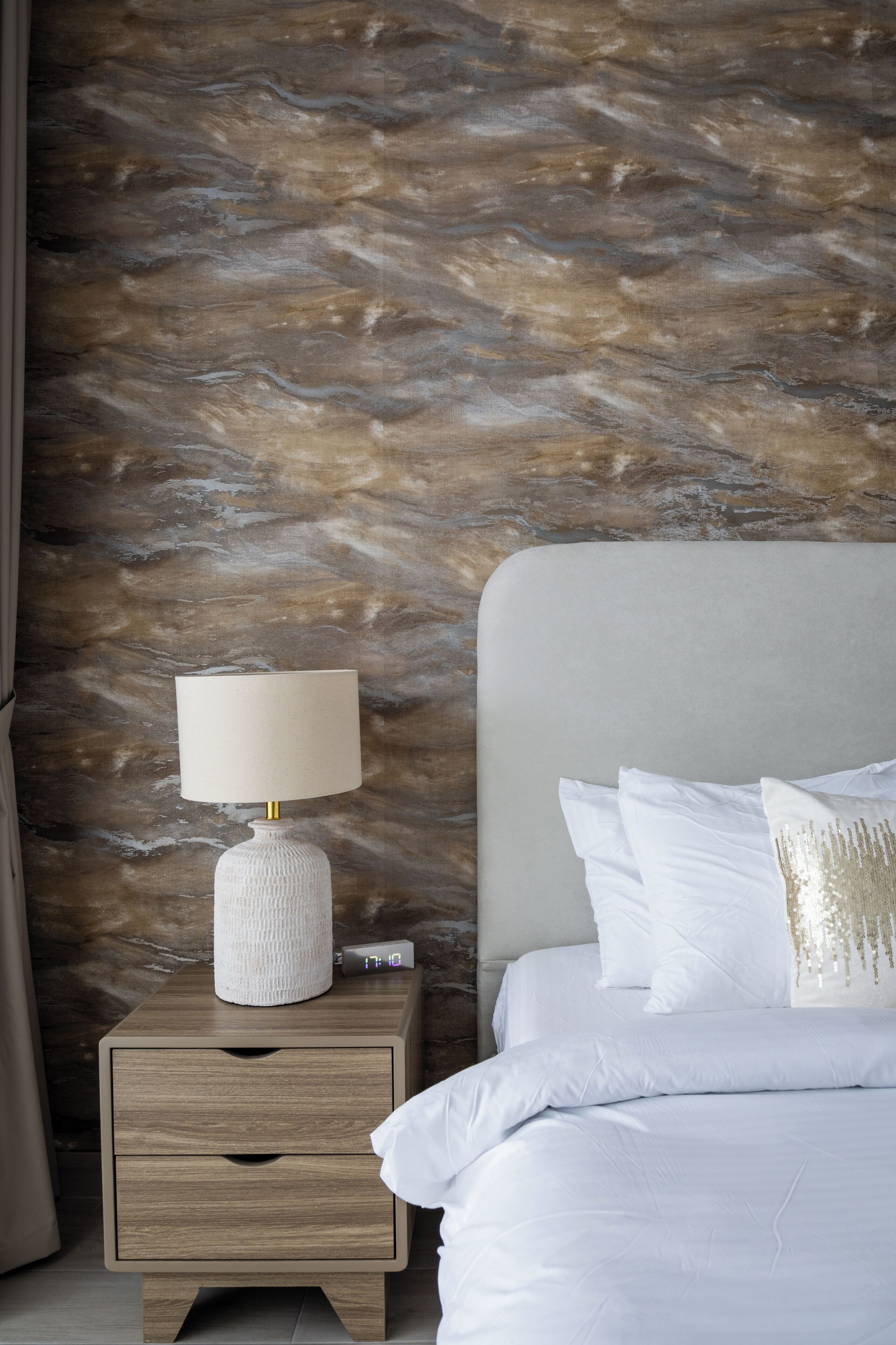 Een net opgemaakt bed met witte lakens en decoratieve kussens, een bedlampje op een houten nachtkastje, en een gestructureerde wand achtergrond.