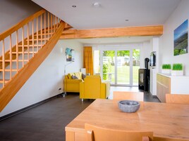 Una sala de estar moderna con una escalera de madera, un sofá amarillo, una mesa de comedor con un tazón y una zona de cocina con un televisor y plantas.