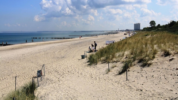 A wide sandy beach with a clear sky and a few clouds. There are people walking and sitting on the beach, and a building is visible in the distance. - Appartementhaus Calmsailing, Borgerende (Boergerende-Rethwisch)