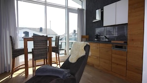 A modern living room with a sofa, a dining table with chairs, a kitchen area, and large windows offering a view of the outside. - Appartementhaus Calmsailing, Borgerende (Boergerende-Rethwisch)