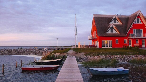 Un muelle de madera que lleva a un edificio rojo con techo pronunciado, barcos amarrados al final y una costa rocosa al fondo.