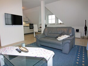 Ein Wohnzimmer mit einer grauen Couch, einem kleinen Tisch mit Fernbedienung und einem an der Wand befestigten Fernseher.