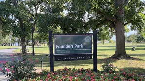 Cartel de Founders Park en Alexandria, con un camino pavimentado y arbustos florecientes al frente.