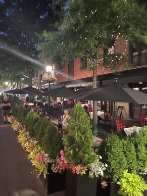 Un área de comedor al aire libre con mesas y sillas, rodeada de plantas en macetas y un edificio de ladrillo al fondo.