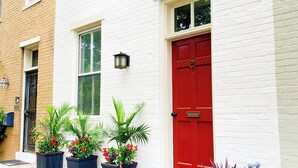 Un edificio de ladrillo blanco con una puerta roja, plantas en macetas y un sendero de ladrillos.